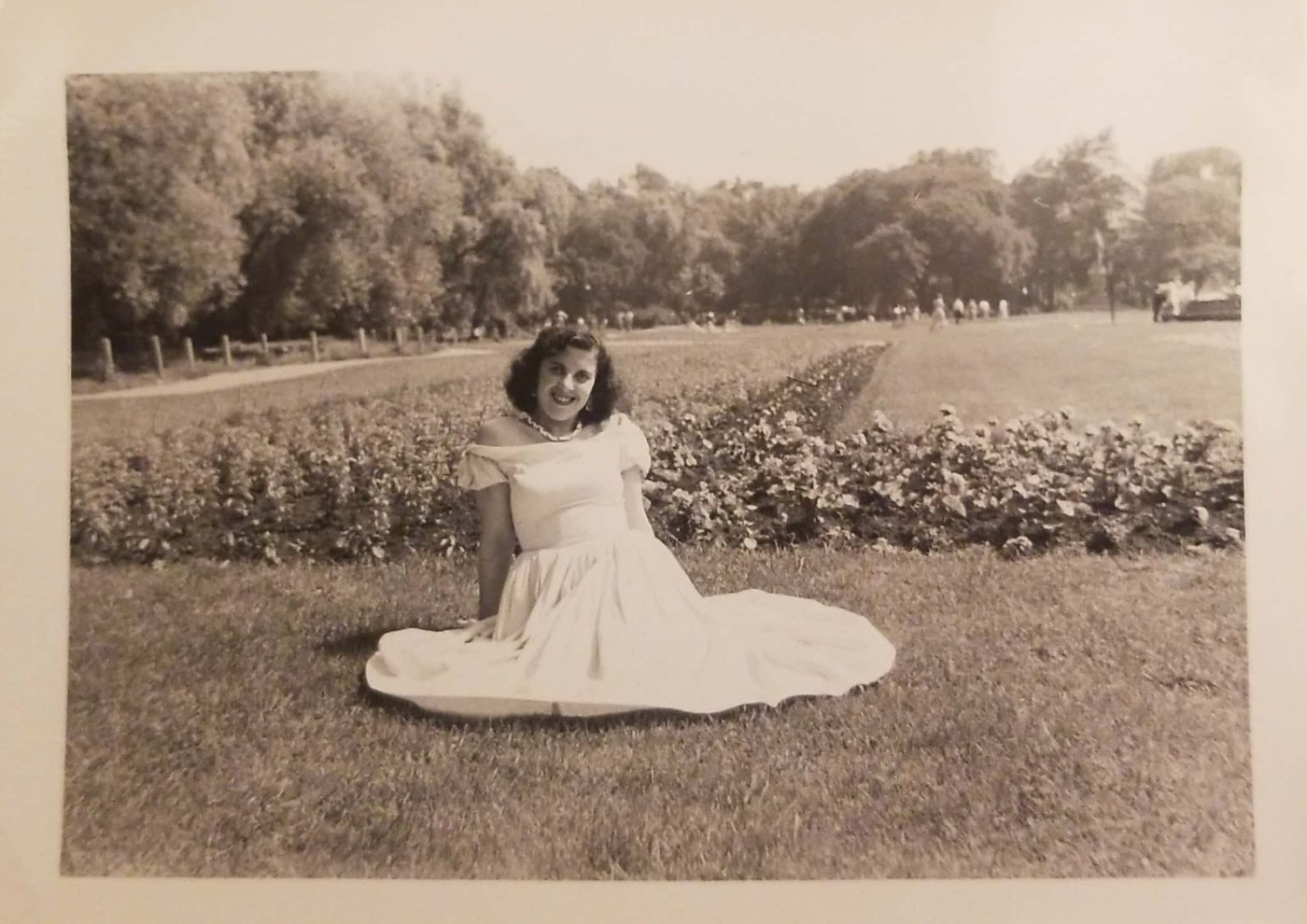 Helen as a young woman in a white off-shoulder gown, smiling, sitting on the grass of a park